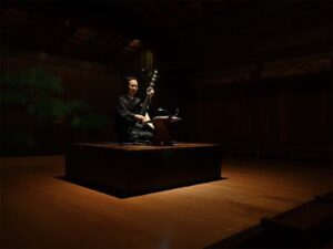 能舞台でスポットライトを浴びながら琵琶を演奏する演奏者。